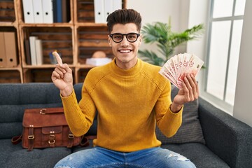 Young hispanic man working at therapy office holding shekels smiling happy pointing with hand and finger to the side