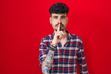 Young hispanic man with beard standing over red background asking to be quiet with finger on lips. silence and secret concept.