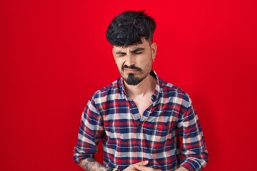 Young hispanic man with beard standing over red background with hand on stomach because indigestion, painful illness feeling unwell. ache concept.
