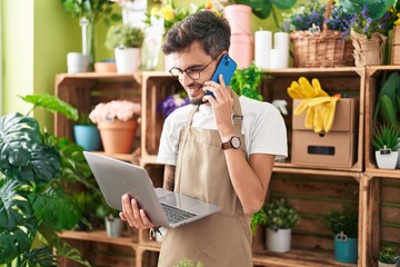 Young hispanic man florist talking on smartphone using laptop at flower shop