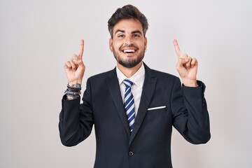 Young hispanic man with tattoos wearing business suit and tie smiling amazed and surprised and pointing up with fingers and raised arms.