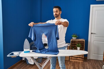 Young hispanic man with beard ironing holding burned iron shirt at laundry room skeptic and...