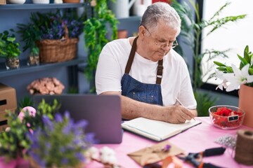 Middle age grey-haired man florist writing on notebook using laptop at florist