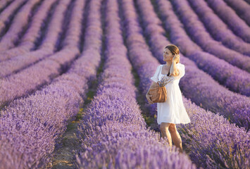 Full body portrait of young brunette woman from in casual white cotton dress enjoying beautiful lavender field and holding woven rattan fashion bag at sunny day