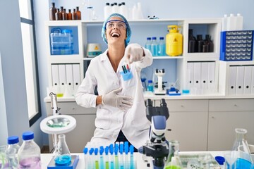 Brunette woman working at scientist laboratory laughing at you, pointing finger to the camera with hand over body, shame expression