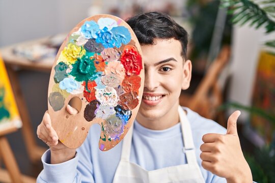Non binary person holding palette close to face smiling happy and positive, thumb up doing excellent and approval sign