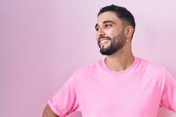 Hispanic young man standing over pink background looking away to side with smile on face, natural...