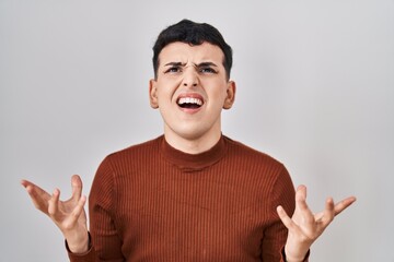 Non binary person wearing make up standing over isolated background crazy and mad shouting and yelling with aggressive expression and arms raised. frustration concept.