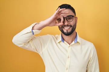 Hispanic young man wearing business clothes and glasses doing ok gesture with hand smiling, eye looking through fingers with happy face.