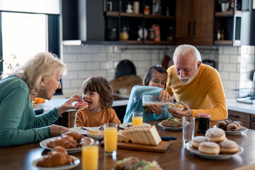 Happy kids enjoying delicious breakfast with caring proud grandparents. Little girl pouring milk into cornflakes cereals with grandpas help while grandma is feeding her little brother with donut. - Powered by Adobe