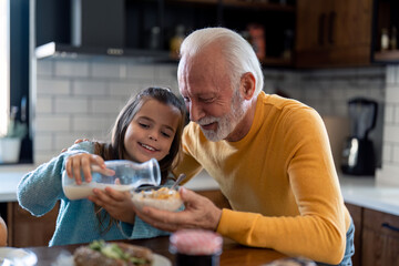 Happy little girl with senior grandfather preparing morning meal in the kitchen at home, adding...