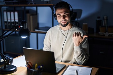 Young handsome man working using computer laptop at night smiling with happy face looking and pointing to the side with thumb up.