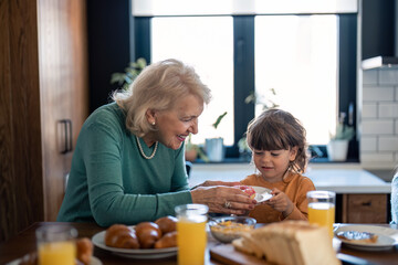 Smiling senior woman proud grandmother giving donut to her adorable small grandson. Lovely small boy having donut for breakfast at grandmas home.