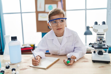 Adorable hispanic boy student writing notes holding molecules at laboratory classroom