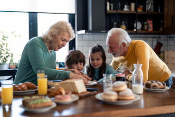 Grandchildren adorable small boy and girl at breakfast with caring grandparents in the kitchen at...