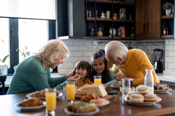 Happy seniors having breakfast at kitchen table with grandkids at home, looking at them with love...