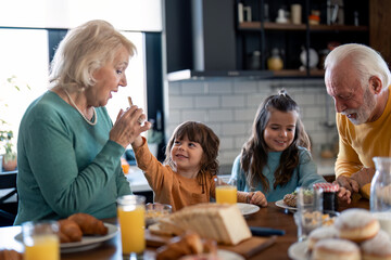 Cute small boy feeding his grandma offering her delicious food during breakfast at home while his sister and grandpa are picking homemade sandwiches. Kids with seniors having morning meal.