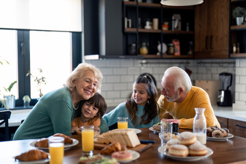 Two cheerful seniors bonding and embracing with grandchildren during breakfast time at kitchen...