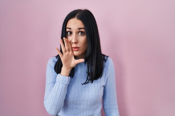 Hispanic woman standing over pink background hand on mouth telling secret rumor, whispering malicious talk conversation