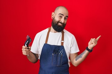 Young hispanic man with beard and tattoos wearing barber apron holding razor with a big smile on face, pointing with hand finger to the side looking at the camera.
