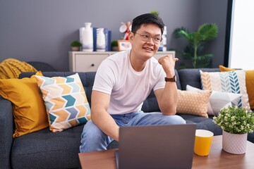 Young asian man using laptop at home sitting on the sofa smiling with happy face looking and pointing to the side with thumb up.