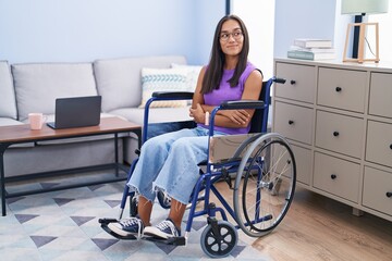 Young hispanic woman sitting on wheelchair at home smiling looking to the side and staring away...