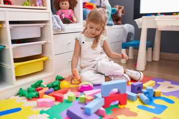 Adorable blonde girl playing with construction blocks sitting on floor at kindergarten