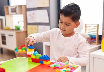 Adorable hispanic boy playing with construction blocks sitting on table at kindergarten