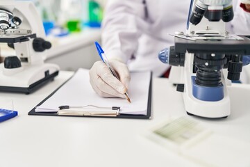 Middle age hispanic woman wearing scientist uniform using microscope write on document at laboratory