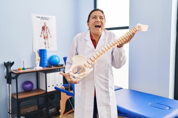 Naklejka na ściany i meble Middle age hispanic woman holding anatomical model of spinal column angry and mad screaming frustrated and furious, shouting with anger looking up.