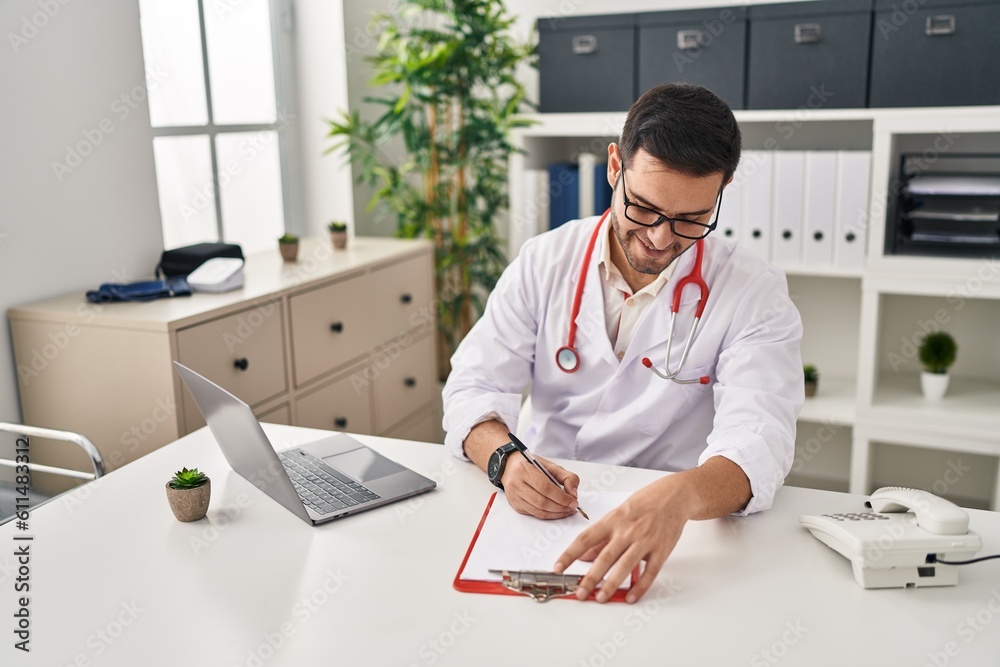 Wall mural Young hispanic man wearing doctor uniform writing report working at clinic