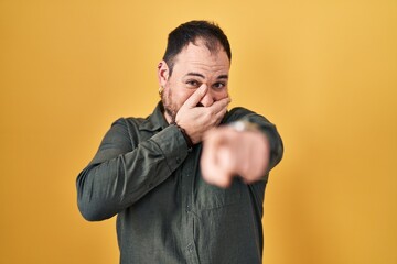 Plus size hispanic man with beard standing over yellow background laughing at you, pointing finger to the camera with hand over mouth, shame expression