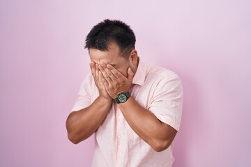 Chinese young man standing over pink background with sad expression covering face with hands while crying. depression concept.