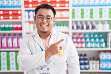 Chinese young man working at pharmacy drugstore cheerful with a smile on face pointing with hand and finger up to the side with happy and natural expression