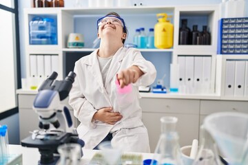 Hispanic girl with down syndrome working at scientist laboratory laughing at you, pointing finger to the camera with hand over body, shame expression