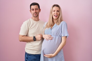 Young couple expecting a baby standing over pink background winking looking at the camera with sexy expression, cheerful and happy face.