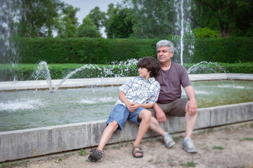 Fototapeta na wymiar Dad and son are relaxing in the city park near fountains. Family have fun. Happy fathers day.