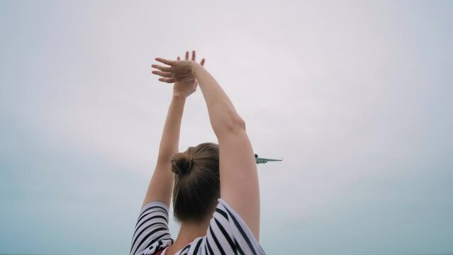 Happy woman waves arms, meets the arriving passenger airplane, airliner against the cloudy gray sky - back view, wide angle view. Positive, vacation, cheering and meeting concept