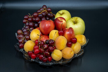 A plate of fruits consisting of grapes, peaches, apricots, cherries and apples on a black background