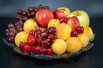 A plate of fruits consisting of grapes, peaches, apricots, cherries and apples on a black background