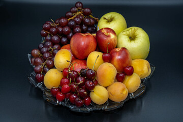 A plate of fruits consisting of grapes, peaches, apricots, cherries and apples on a black background