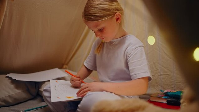 Close-up beautiful creative little girl hiding inside a tent, drawing pictures with colorful pencils