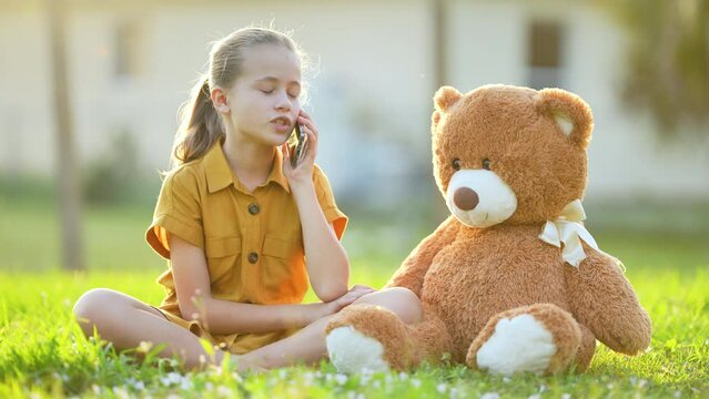 Pretty preteen girl playfully taking with her friend on sell phone sitting outdoors on green grass lawn. Concept of friendship values