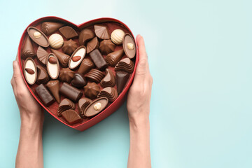 Woman holding heart shaped box with delicious chocolate candies on light blue background, top view....