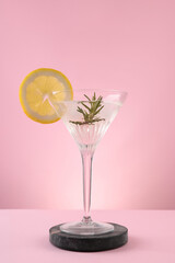 Martini glass of cocktail with lemon slice and rosemary on stand against pink background