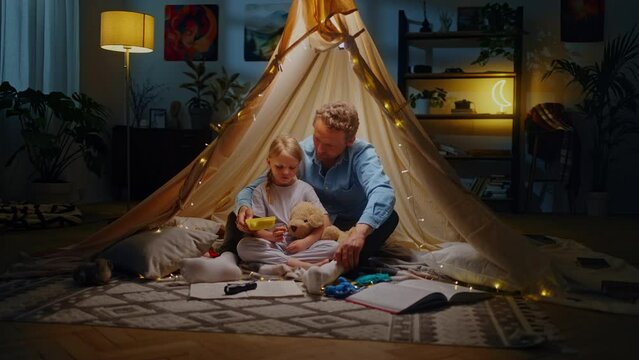 Loving single father embraces his daughter while plays tent on floor, snack together inside a tent