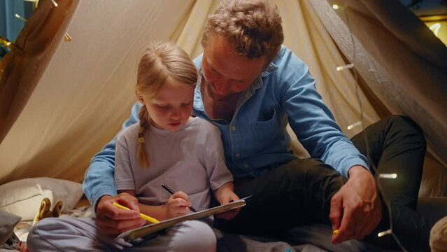 Happy dad and daughter playing drawing pictures with color pencils inside a play wigwam tent at home