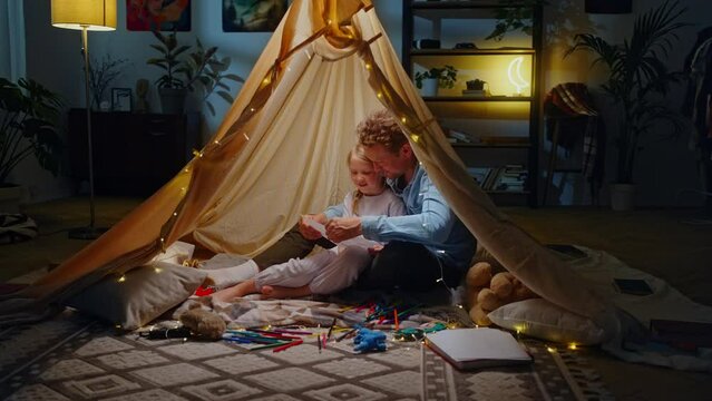 Happy dad hugging his daughter, admiring her drawn picture while sitting inside a wigwam at home