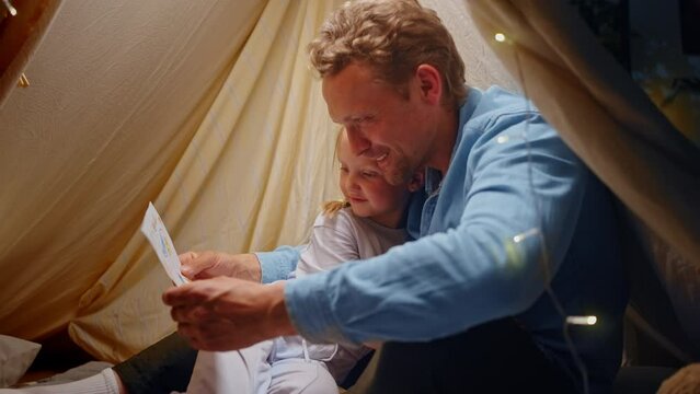 Loving dad, hugs his daughter admiring her drawing, sitting together inside tent on floor at home