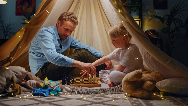 Slow motion. Happy father teaches her daughter to play chess, sitting together in a wigwam tent
